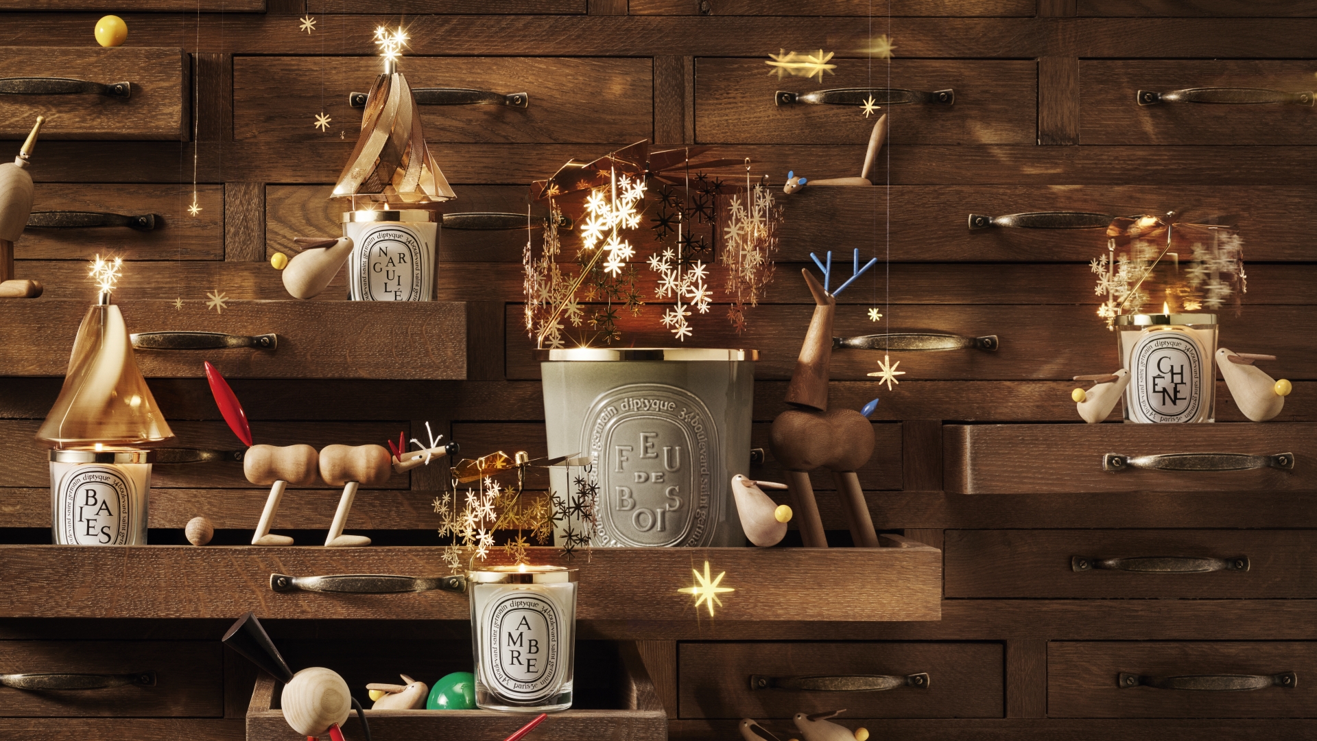 A flat lay of Diptyque's 2026 holiday collection items, featuring green glass scented candles, festive packaging, and dark wood oval labels.